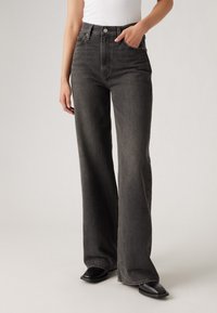 Jean large en denim noir, doté d'une taille haute, de poches avant et d'une finition épurée avec un matériel minimal. La texture du tissu semble robuste.