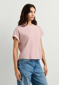 Young woman with long dark hair wearing a light pink short-sleeve t-shirt and blue denim jeans, standing against a plain background.