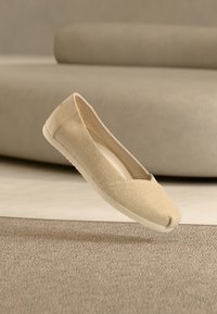 Μπεζ επίπεδο παπούτσι τύπου slip-on με υφασμάτινο επάνω μέρος και λευκή σόλα, τοποθετημένο στον αέρα πάνω από δάπεδο με χαλί, με έναν μπεζ καναπέ στο φόντο.