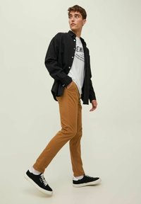Brown trousers, black button-up shirt, and white graphic t-shirt with black text. Model wears black sneakers with white accents.