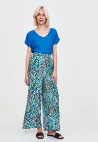 Blaues V-Ausschnitt-Oberteil mit kurzen Ärmeln, kombiniert mit weit geschnittenen Hosen mit einem mehrfarbigen Tierdruck. Flache schwarze Sandalen runden das Outfit ab.