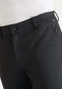 Zwarte broek met een subtiele textuurpatroon, voortassen en een knoopsluiting. Inclusief een kleine driekleurige accenten bij de tailleband.
