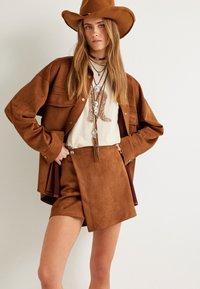 Veste en suède marron et short avec un t-shirt graphique crème. Les accessoires comprennent des colliers superposés et un chapeau à larges bords marron.