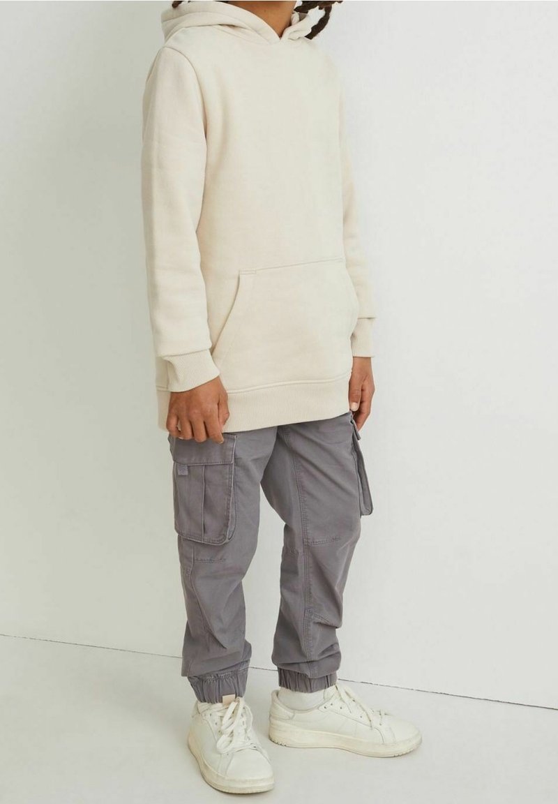 Enfant portant un sweat à capuche crème surdimensionné, un pantalon cargo gris avec poches latérales, et des baskets blanches, debout contre un mur clair uni.