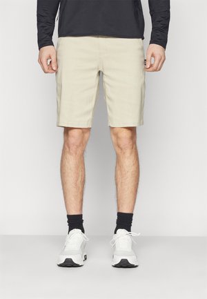 Mann trägt beige Shorts, schwarzen langärmligen Oberteil, schwarze Socken und weiße Turnschuhe und steht vor einem schlichten hellen Hintergrund.