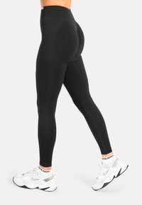 Svarta högmidjade leggings med en figurnära design, tillverkade av stretchigt material med texturerade detaljer längs sidorna. Vita sneakers syns.