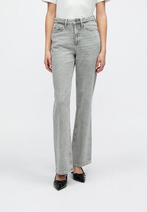 Femme portant un jean droit taille haute gris clair et des ballerines noires à bout pointu, debout devant un fond blanc uni.