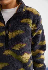 Polarowy pulower w czarno-zielono-fioletowym wzorze kamuflażu, z półzamkiem oraz kieszenią na zamek na piersi z logo.