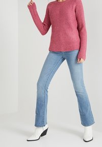 Ribbed, maroon sweater with long sleeves, paired with light blue flared jeans. Model wearing black and white ankle boots. Simple background.