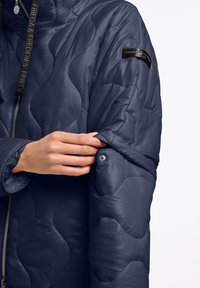 Gevoerd blauw pufferjack met een gestructureerd oppervlak, ritsluiting en een gelabelde mouwpatch. Heeft een knopdetail op de mouw.