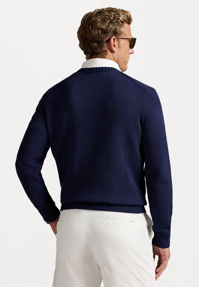 Maglione in maglia blu navy con scollo a girocollo classico, polsini e orlo a costine. Indossato sopra una camicia bianca a colletto, abbinato a pantaloni chiari.