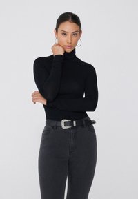 Un haut col roulé noir à manches longues, associé à un jean sombre taille haute et une ceinture noire avec une boucle en métal argenté.