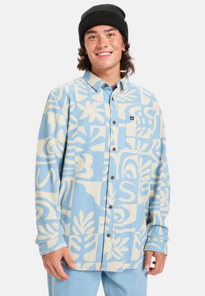Button-up shirt in light blue with large cream abstract patterns, featuring a chest pocket and brown buttons. Worn over light jeans.