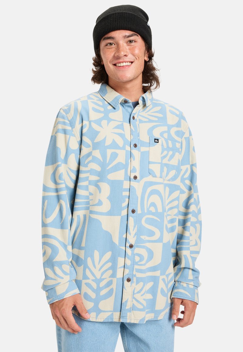 Camisa de botón en azul claro con grandes patrones abstractos en color crema, con un bolsillo en el pecho y botones marrones. Lleva puesta sobre jeans claros.