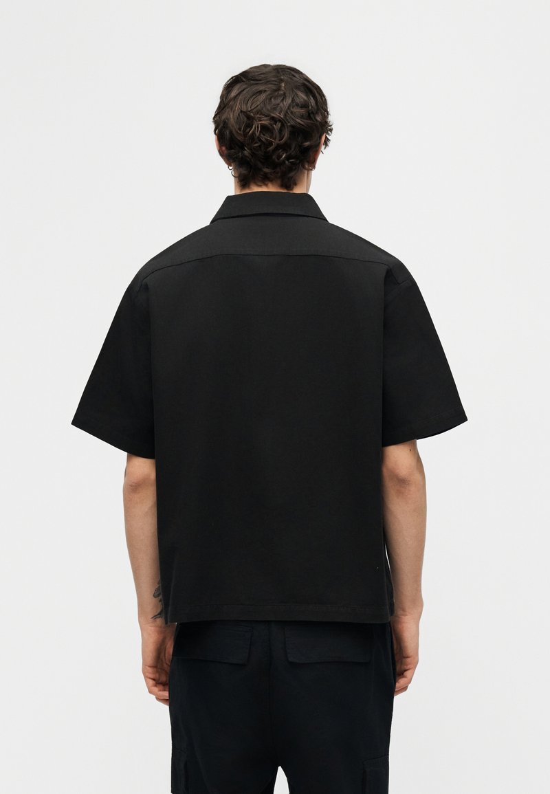 Uomo con capelli ricci che indossa una maglietta nera a maniche corte e pantaloni neri, in piedi con la schiena rivolta verso lo spettatore, davanti a uno sfondo chiaro e uniforme.