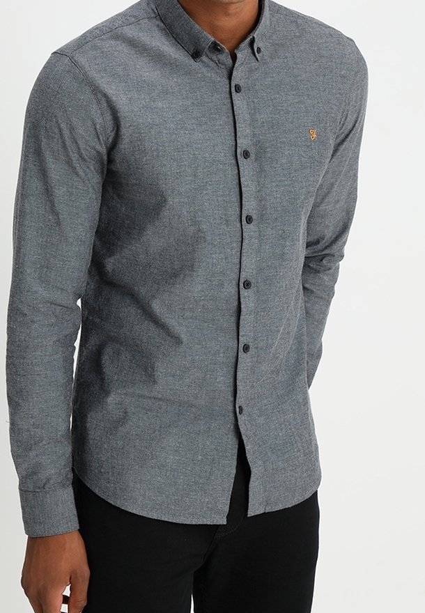 Homme portant une chemise grise cintrée à manches longues boutonnée avec un pantalon noir sur un fond clair uni.