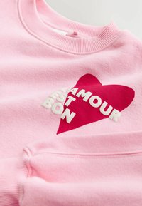 Sudadera rosa hecha de tela suave con un gran gráfico de corazón rojo y texto blanco "L’AMOUR EST BON" en el frente. Puños acanalados.