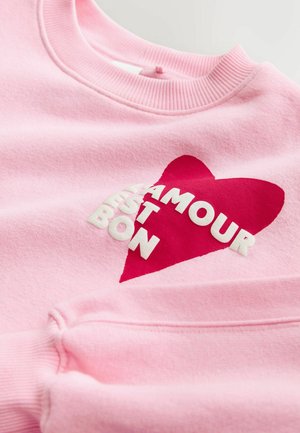 Lyserød sweatshirt lavet af blødt stof med stort rødt hjerteprint og hvid tekst "L’AMOUR EST BON" på forsiden. Ribstrikkede manchetter.