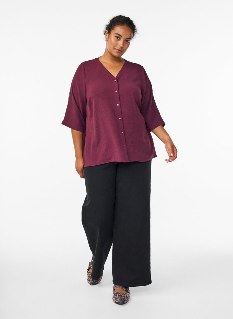 Woman wearing a loose maroon buttoned top, wide-leg black pants, and leopard print shoes walking against a plain white background.