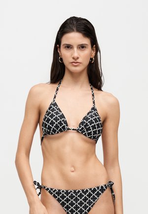 Kobieta z długimi ciemnymi włosami, ubraną w czarno-biały wzorzysty bikini typu halter i dopasowane majtki z wiązaniem po bokach, stojąca przodem na jednolitym tle.