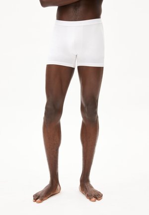 Person steht barfuß in schlichten weißen Boxershorts vor einem einfarbigen hellen Hintergrund mit verschränkten Armen.