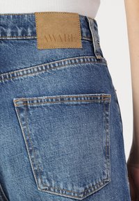 Jeans de mezclilla azul presentan un parche de cuero marrón con la etiqueta "AWARE" en la cintura y bolsillos traseros cosidos con detalles de hilo naranja.