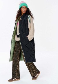 Schwarzige gesteppte lange Weste mit grünem Innenfutter, getragen über einem beigen gemusterten Pullover und Babyschuhen aus Leopardenmuster, kombiniert mit braunen Sneakers und einer türkisfarbenen Mütze.