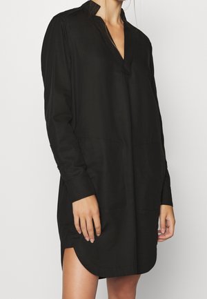 Femme portant une robe chemise noire à manches longues, ample, avec un col en V et une fente latérale, sur un fond uni.