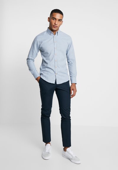 Camisa azul clara com botões, de mangas longas, com colarinho pontudo e botões bege, combinada com calças estreitas cinza-escuro e ténis brancos.