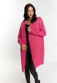 Cárdigan rosa y esponjoso de manga larga, con acentos similares a perlas en los hombros y un corte holgado. Llevado sobre una camiseta y pantalones negros.