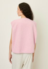 Rozā flīsa veste ar teksturētu virsmu, plašiem roku atverēm un noapaļotu apakšmalu, stila no aizmugures uz neitrāla fona.