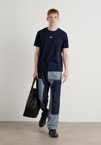 Tricou de culoare bleu marin cu guler rotund și un mic logo, asortat cu pantaloni din denim patita gri și albastru. Modelul poartă o geantă neagră și încălțăminte neagră.
