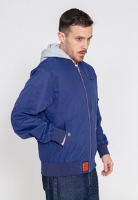 Marine bomberjack met een grijze capuchon, ritsluiting, zijzakken en geribde manchetten. Kenmerkt een oranje label op de voorzoom. Gemaakt van een katoenmix.