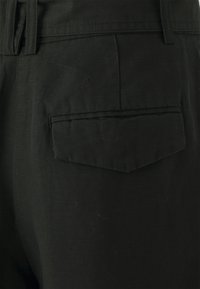 Pantaloni neri con tessuto testurizzato, dotati di una tasca posteriore e passante per cintura. La tasca ha una forma rettangolare con un dettaglio a pattina.