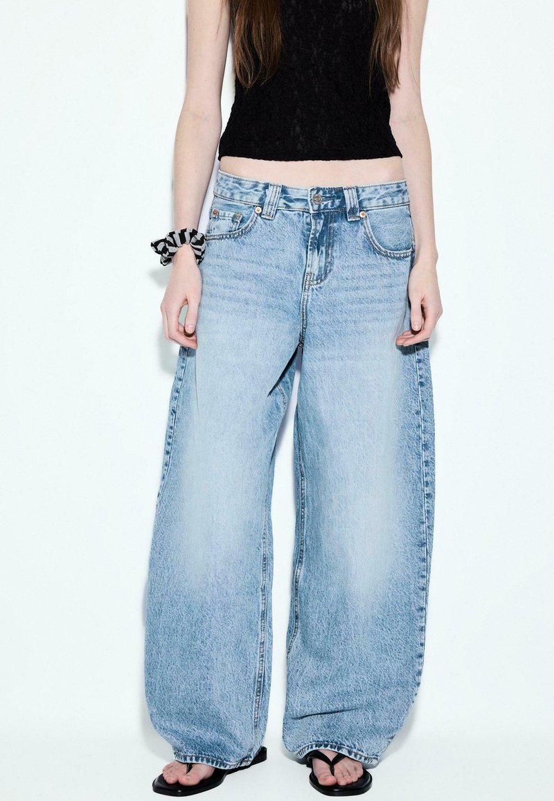 Jeune personne portant un jean large bleu clair, un crop top en dentelle noire, un scrunchie noir et blanc au poignet gauche, et des tongs noires.