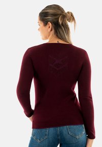 Maglione bordeaux a maniche lunghe con scollatura rotonda e dettaglio del logo testurizzato sulla parte posteriore, abbinato a jeans in denim blu con cuciture decorative.