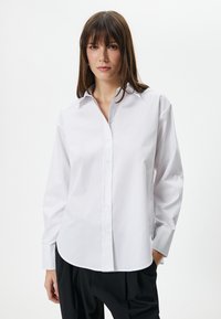 Camicia bianca in cotone con colletto appuntito, maniche lunghe, chiusura con bottoni e orlo arrotondato. Indossata con pantaloni neri. Texture liscia.