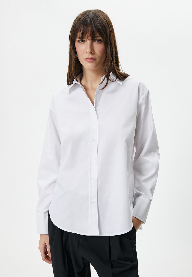 Camicia bianca in cotone con colletto appuntito, maniche lunghe, chiusura con bottoni e orlo arrotondato. Indossata con pantaloni neri. Texture liscia.