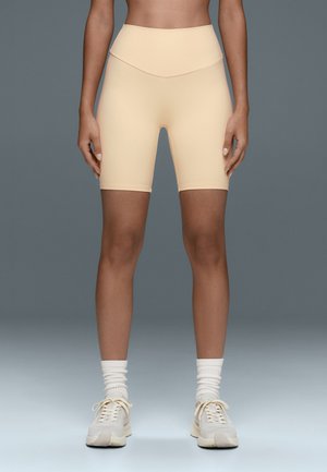 Person, die beige High-Waist-Biker-Shorts, weiße gerippte Socken und beige Sneakers trägt und vor einem einfarbigen grauen Hintergrund steht.