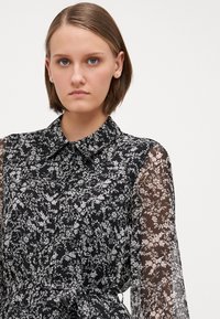 Svart bluse med blomsterdesign, krage, gjennomsiktige lange ermer og knyteliv. Har et delikat, lett stoff.
