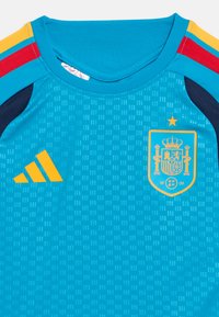 Zils sporta krekls ar dzelteno Adidas logo, Spānijas nacionālās futbola komandas emblēmu un sarkaniem, dzelteniem un zilajiem svītrām uz pleciem.