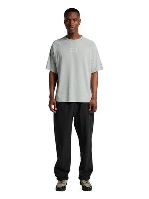 Mand står iført en lysegrå kortærmet skjorte med "223" print, sorte løstsiddende bukser og beige og sorte sneakers på hvid baggrund.