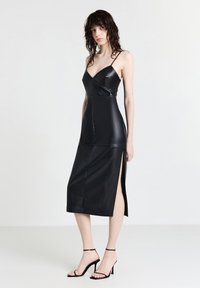 Frau mit nassem Haar, die ein schwarzes Leder-Midikleid mit dünnen Trägern, seitlichem Schlitz und schwarzen hochhackigen Sandalen trägt, steht vor einem weißen Hintergrund.
