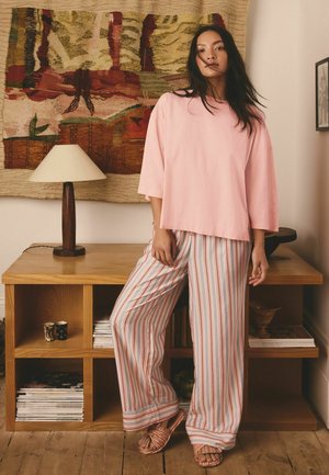 Žena stojí v interiéri a má na sebe voľnú ružovú blúzku a široké pruhované nohavice, s sandálmi, pred drevenou policou a abstraktným nástenným závesom.