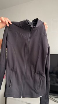 Veste noire à fermeture éclair, en tissu lisse, dotée d'un col montant, de deux poches avant et de coutures décoratives sur les épaules.