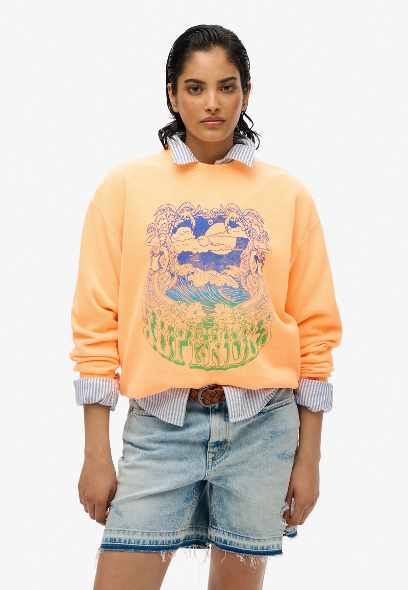 Sudadera naranja con un diseño gráfico azul que presenta olas y nubes. Llevada sobre una camisa de rayas con cuello y combinada con pantalones cortos de mezclilla.