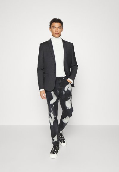 Schwarzes Sakko, weißer Rollkragenpullover und schwarze Jeans mit einem weißen Batikmuster. Die Sneakers sind schwarz mit weißen Sohlen und Schnürsenkeln.