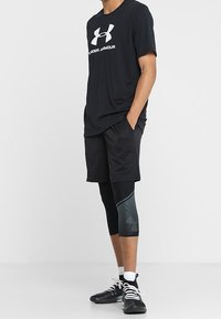Hombre de pie con las manos en los bolsillos, vestido con una camiseta negra de Under Armour, pantalones cortos negros, mallas negras y grises, calcetines blancos y zapatillas negras.