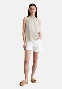 Top halter tan hecho de lino, combinado con pantalones cortos de denim blanco. La modelo lleva sandalias trenzadas con suela de plataforma, de pie contra un fondo neutro.