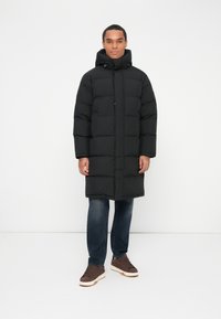 Mand iført en lang sort pufferjakke med hætte, blå jeans og brune sneakers, stående foran en ensfarvet hvid baggrund.